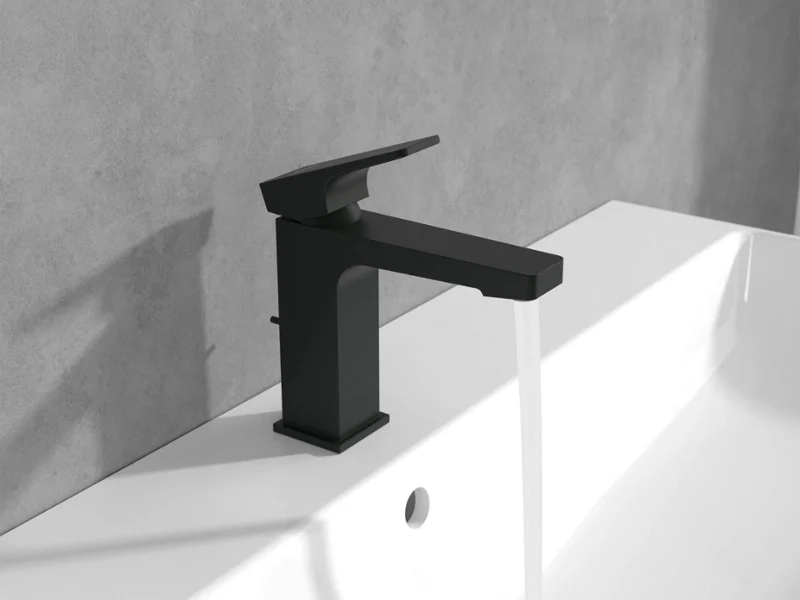 Czarna bateria Architectura Square na białej umywalce Czarna bateria, z której woda spływa do białej umywalki na tle szarej ściany.