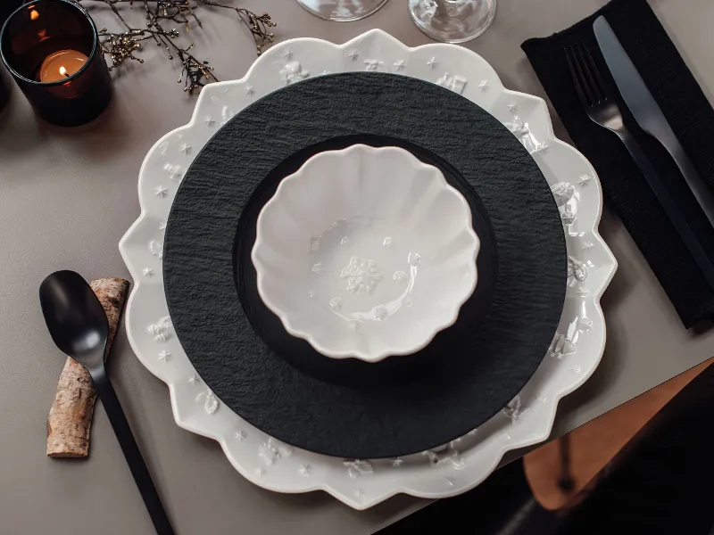 Une table dressée en noir et blanc. On y trouve des assiettes Villeroy & Boch Toy’s Delight Royal Classic avec un bord ondulé et des assiettes rondes, un bol blanc, des couverts de table noirs, une serviette de table et trois bougies noires allumées.