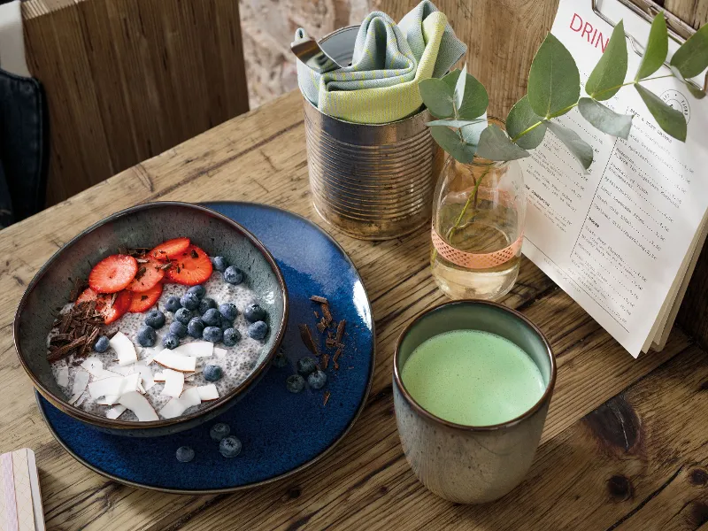 A healthy smoothie bowl on a wooden table in a Lave bowl, a Lave cup of matcha latte, and a menu.