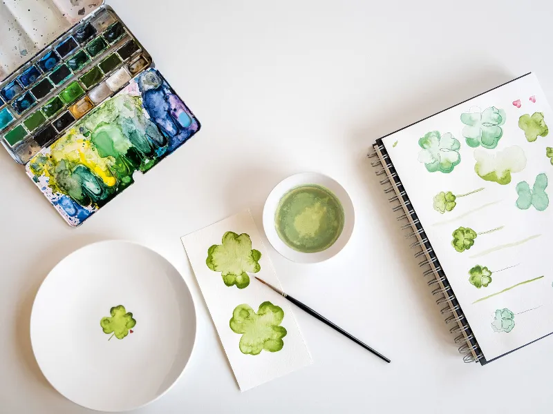 A watercolour set-up with a Villeroy & Boch “With Love" plate and a sketchbook decorated with green shamrock pictures on a white table.