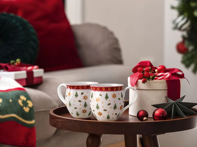 Two Christmas mugs, a gift box, and Christmas decorations on a small table next to a festively decorated living room.
