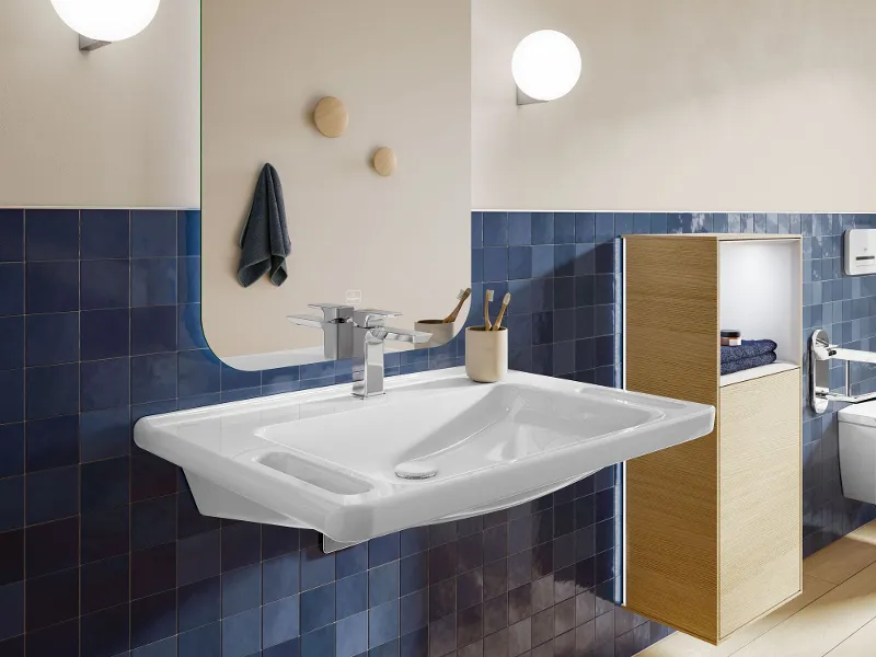 Un moderno bagno senza barriere con un lavabo rettangolare ViCare dotato di maniglie per una maggiore sicurezza, pareti piastrellate blu, un grande specchio, due applique e una mensola in legno.