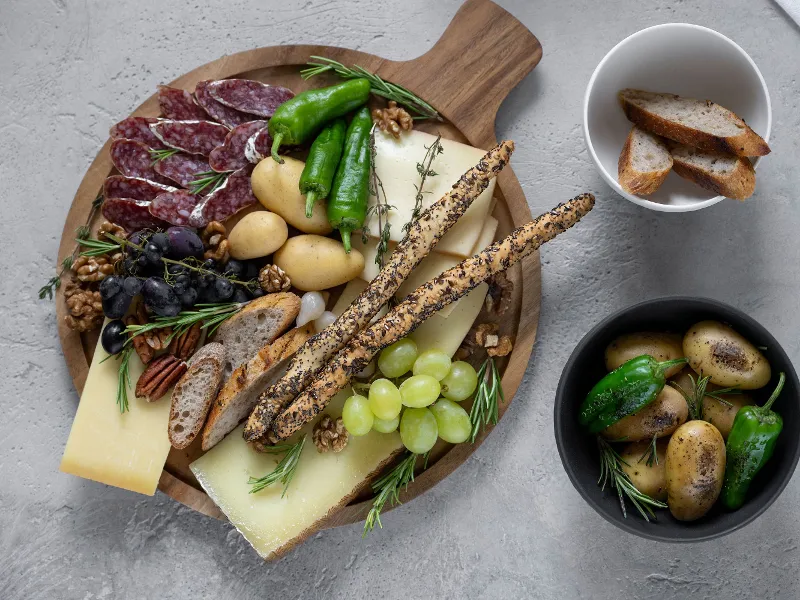 Cheese, salami, grapes, nuts, potatoes, peppers and grissini are elegantly arranged on a Villeroy & Boch Artesano wooden board. Next to this is a bowl of sliced bread and another bowl of potatoes and peppers. Cheese, salami, grapes, nuts, potatoes, peppers and grissini are elegantly arranged. 