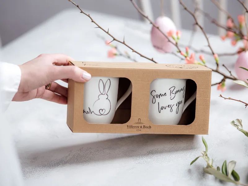 Woman's hand holding a pretty gift box containing 2 cups with a bunny pattern. In the background are Easter branches with pink decorative eggs.