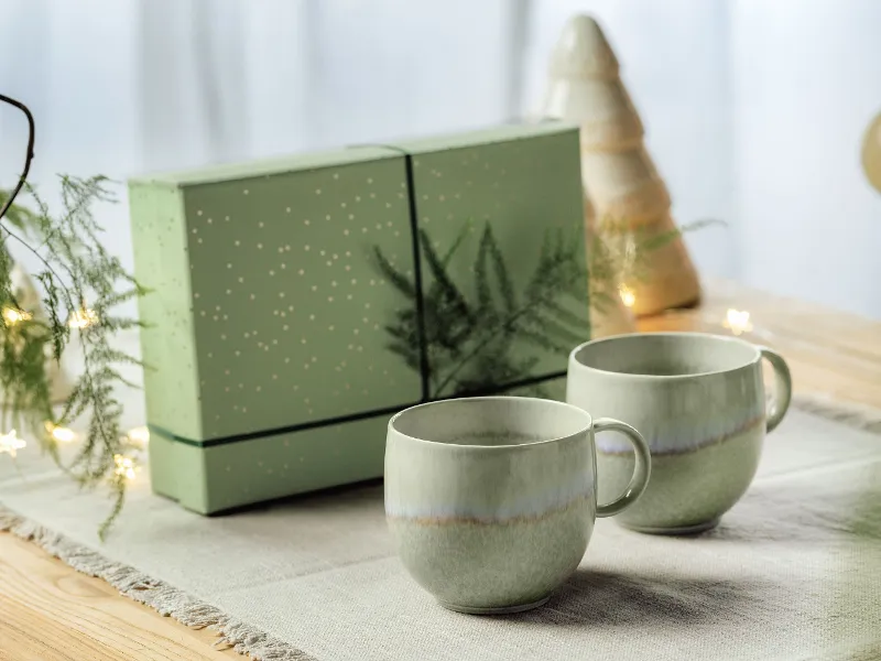 Two green Perlemor cups on a table with a matching green gift box in the background, decorated with a tree and lights.