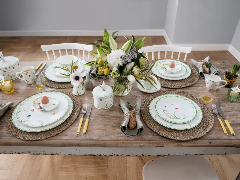 A Villeroy & Boch "Colourful Spring" dinner set for four people consisting of floral-patterned porcelain, cutlery with yellow handles, woven placemats and a floral arrangement. Each place setting includes an egg in a decorative cup.