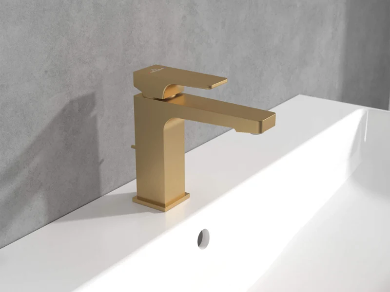 Złota bateria Architectura Square na białej umywalce Nowoczesna złota bateria zamontowana na białej umywalce, na tle szarej ściany.