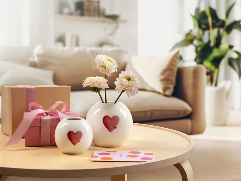 Due vasi bianchi con motivi a cuori rossi di with love, una confezione regalo rosa e un biglietto su un tavolino rotondo in un accogliente salotto.