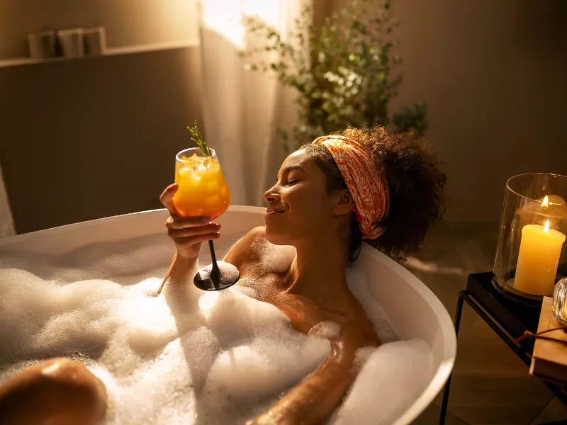 A woman with a drink in her hand lying in a bath in candlelight.