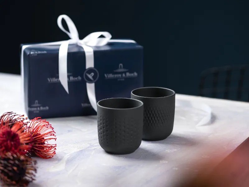 Two black collier cups on a table with a gift box, next to a red and black flower.