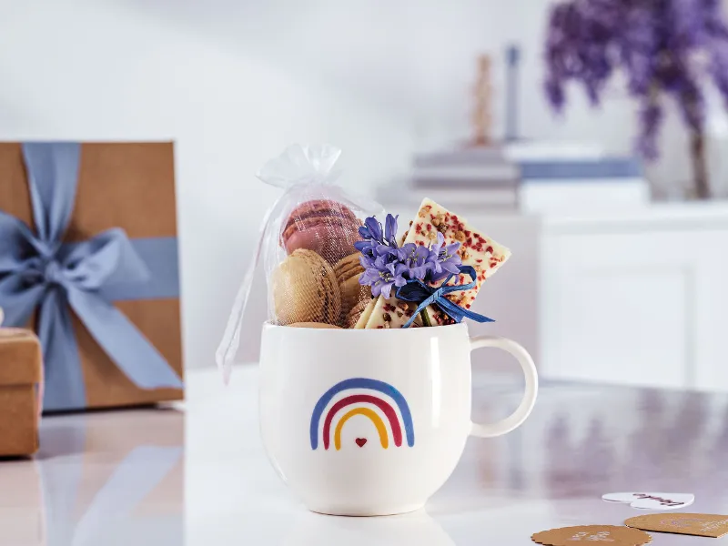 A Villeroy & Boch "With Love" cup with a rainbow design, filled with macarons, a bar of chocolate and purple flowers on a white surface with gift boxes and books in the background.