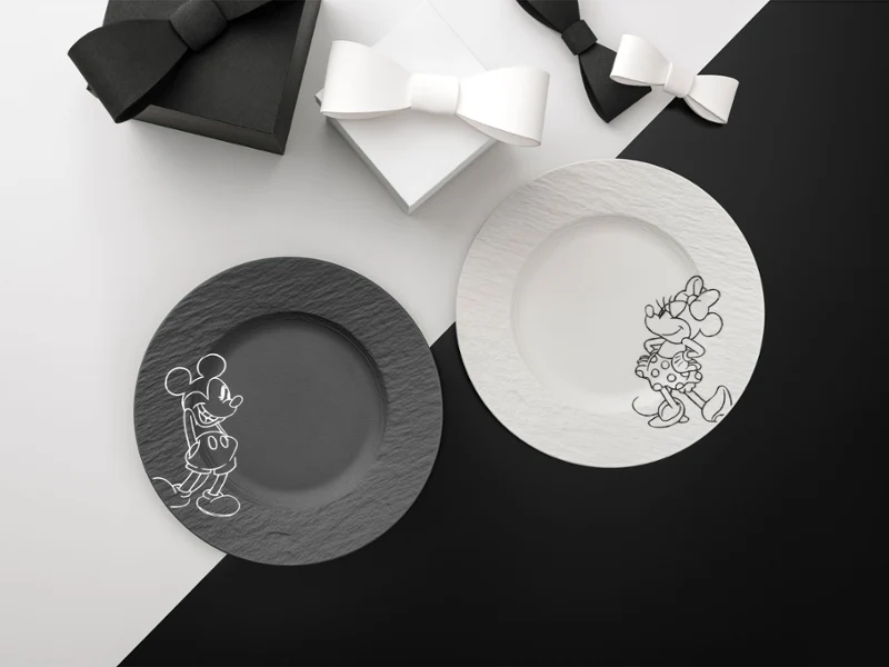Manufacture Rock Mickey plato negro y blanco sobre fondo blanco y negro Dos platos de la colección Manufacture Mickey, uno negro y otro blanco, sobre un fondo dividido en blanco y negro con dos cajas de regalo con lazo.