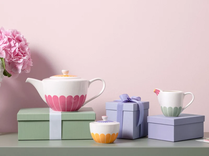 Teapot, milk jug, and sugar bowl with colorful floral patterns from the Fleur collection and pastel-colored gift boxes are arranged on a green surface with pink and white flowers in the background.