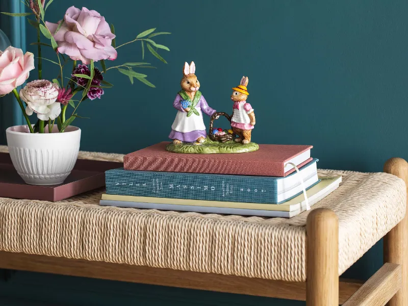 To dekorative harefigurer fra kolleksjonen Bunny Tales fra Villeroy & Boch oppå stablede bøker på en flettet benk, ved siden av en vase med ranunkler og roser.