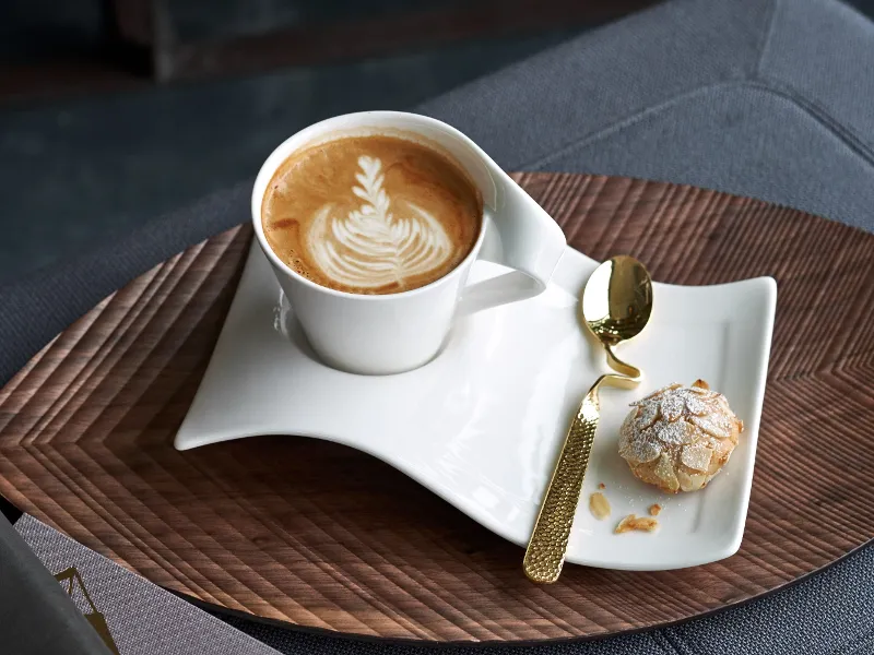 A Villeroy & Boch NewWave latte cup with latte art on a wavy white saucer is combined with a small cookie and a golden spoon, all beautifully arranged on a wooden tray.