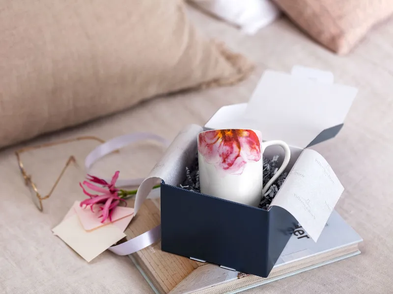 An open Villeroy & Boch gift box with a Rose Garden cup, pink flowers and a note on a wooden tray, with soft cushions in the background.