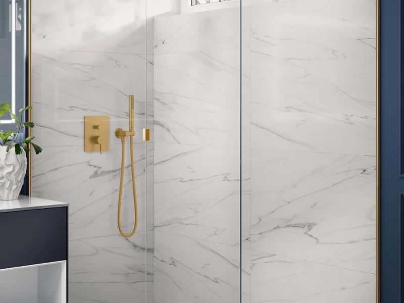 Golden Universal Shower fitting in a marble-look shower Modern glass shower with white marble-look walls, golden fittings and a window letting in natural light.
