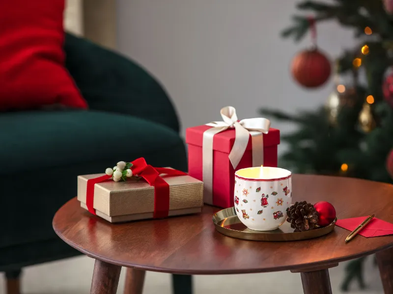 Un tavolo di legno con due regali incartati, una candela accesa in una tazza festiva, pigne e una busta rossa vicino a un albero di Natale decorato e una sedia verde.
