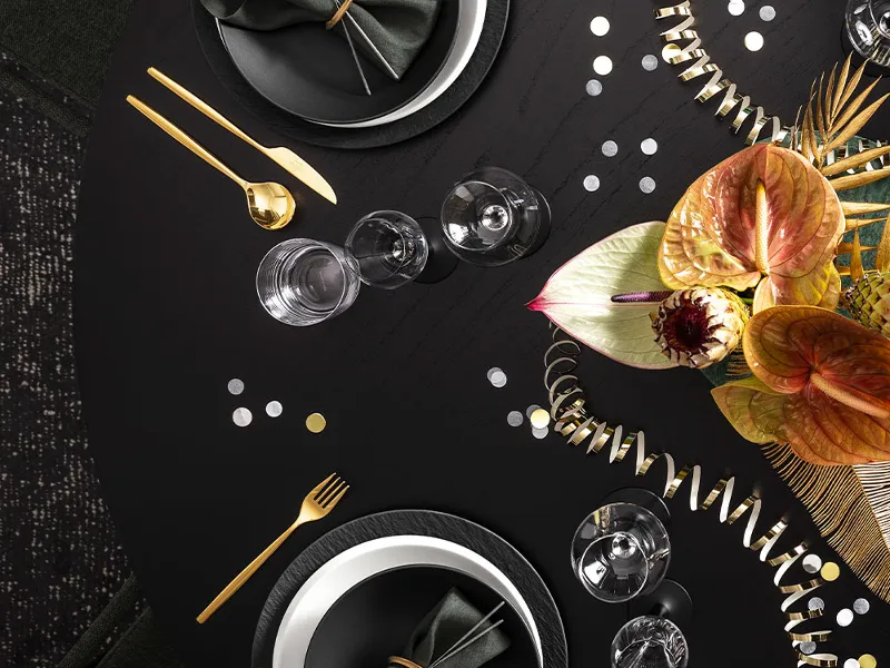 Une table ronde pour quatre personnes avec des assiettes noires Manufacture de Villeroy & Boch, des couverts de table dorés et des serviettes vertes. Une composition florale avec des feuilles orange et vertes en décore le centre.