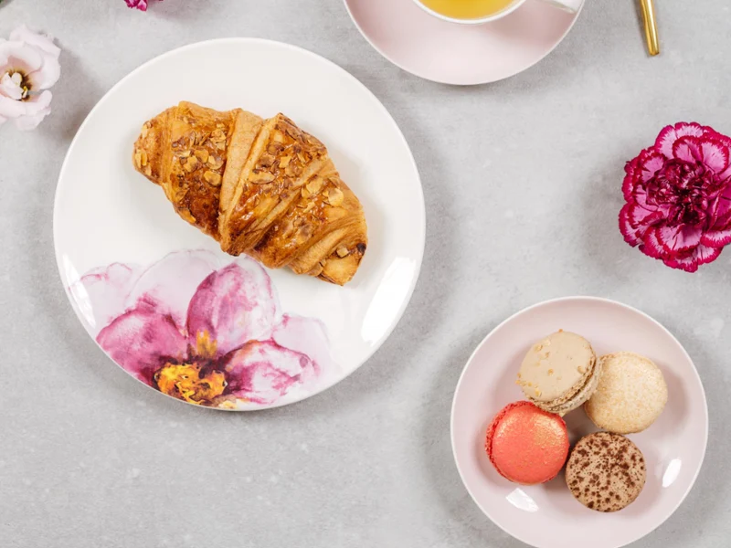 An elegant afternoon tea setting with a croissant, macarons, a cup of tea and fresh flowers with Rose Garden crockery on a marble surface.