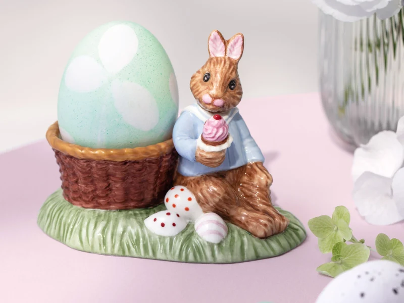 Ceramic Annual Easter Edition 2026 bunny figurine in a blue jacket sits beside a large pastel egg in a basket on a grassy base with small eggs.