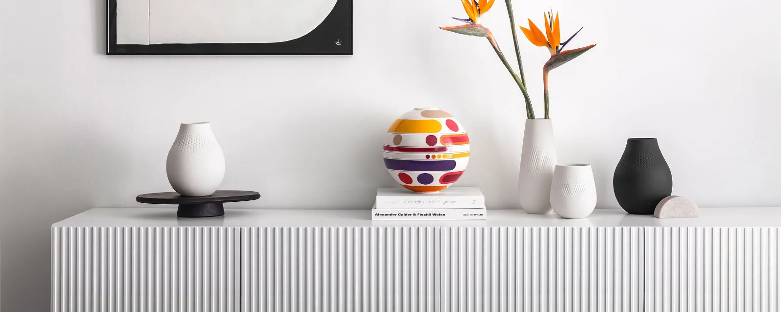 A white sideboard with vases, the colorful La Boule Miami and books underneath, next to a bird of paradise flower arrangement, with geometric wall art in the background.