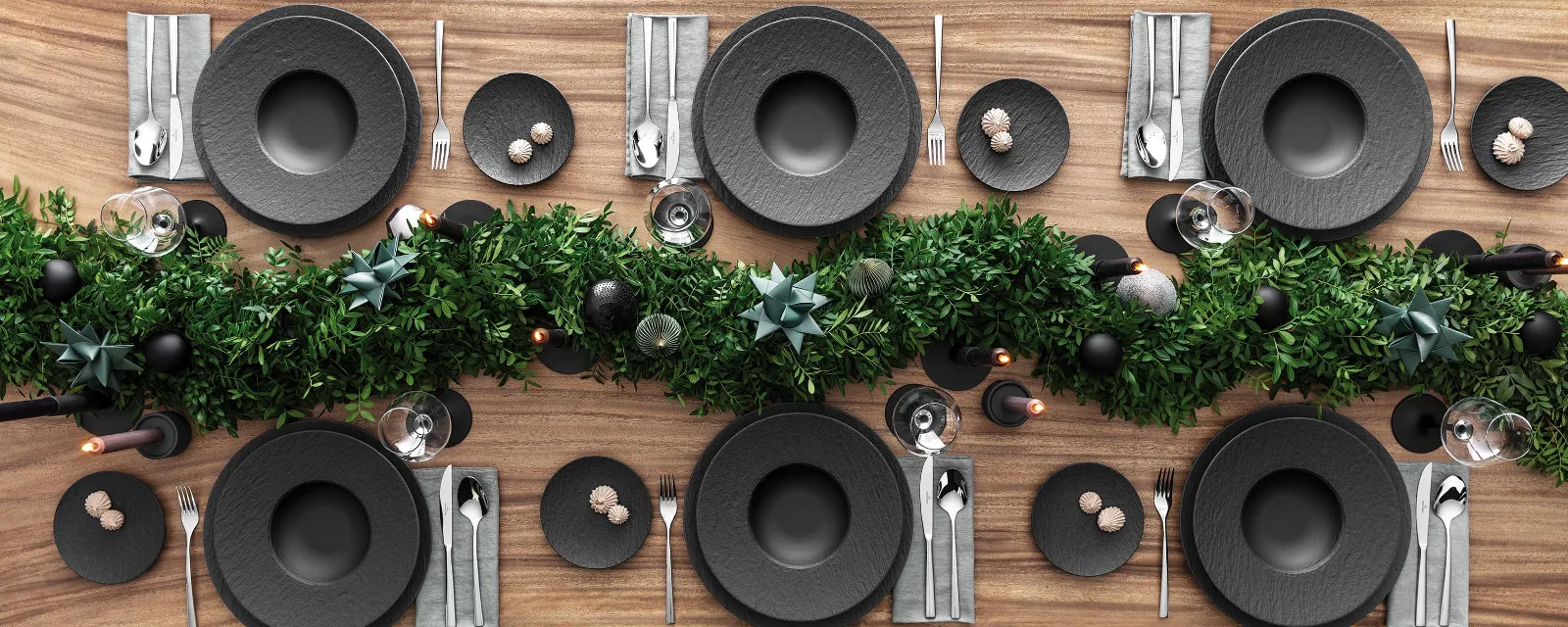 A top view of a dining table set for Christmas with black plates from the Manufacture range by Villeroy & Boch and complemented by Piedmont cutlery. It is also decorated with a centerpiece of green foliage and candles on a wooden surface.