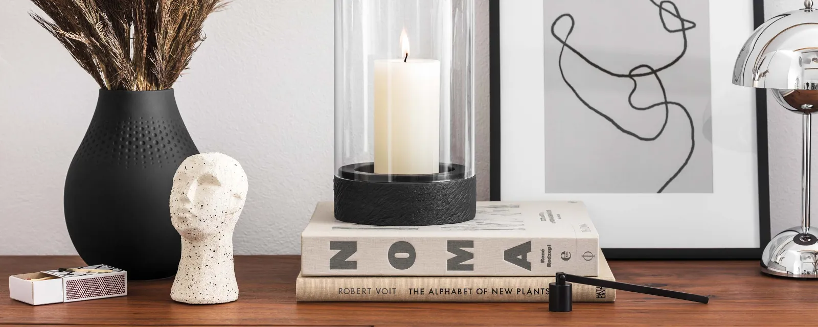 A modern highboard with decorative objects, including a vase with dried plants, a black Manufacture hurricane lamp, a sculpture, stacked books and a framed abstract artwork.