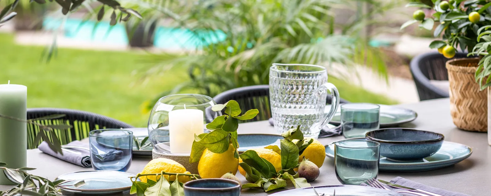 Outdoor dining table with Lave crockery, glasses, candles and fresh citrus fruits as a centrepiece with a view of a pool.