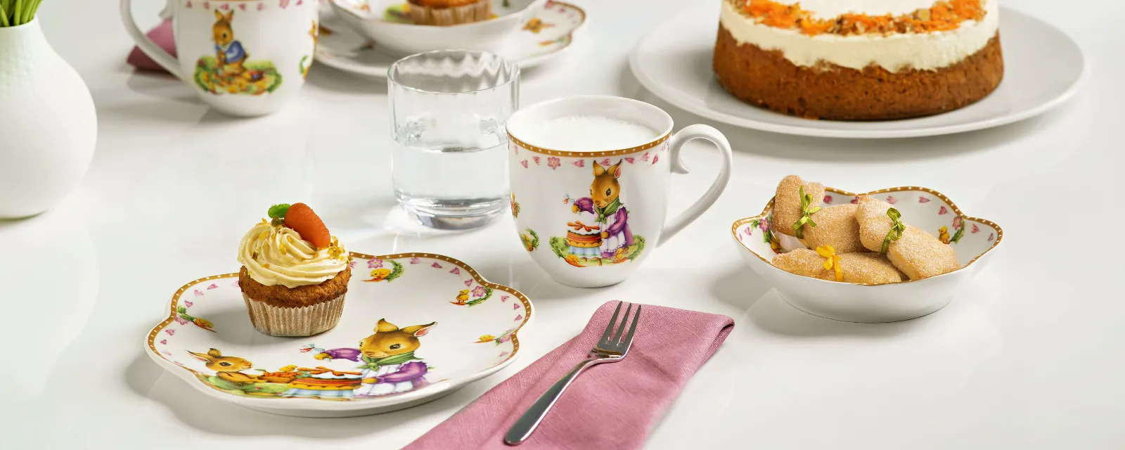 A table with tea cups and plates from the Annual Easter Edition 2024 collection with various pastries and a sponge cake, all adorned with spring decorations.