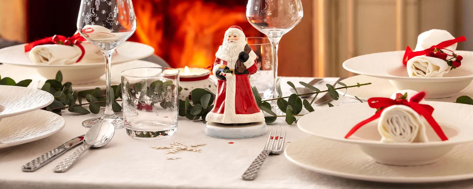 Festive table setting with white Manufacture plates, rolled napkins, two wine glasses, Boston flatware, and a small Santa Claus figure. A fireplace with a burning fire can be seen in the background.