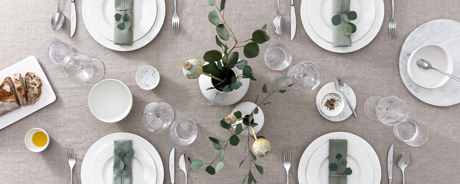 A table setting with a beige tablecloth, white Manufacture plates, cutlery, wine glasses and a flower arrangement