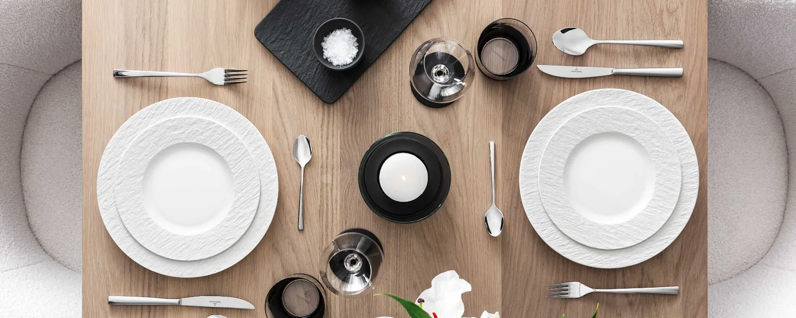 Elegant dining table with white Manufacture plates, Piemont cutlery and decorative flowers on a wooden surface.