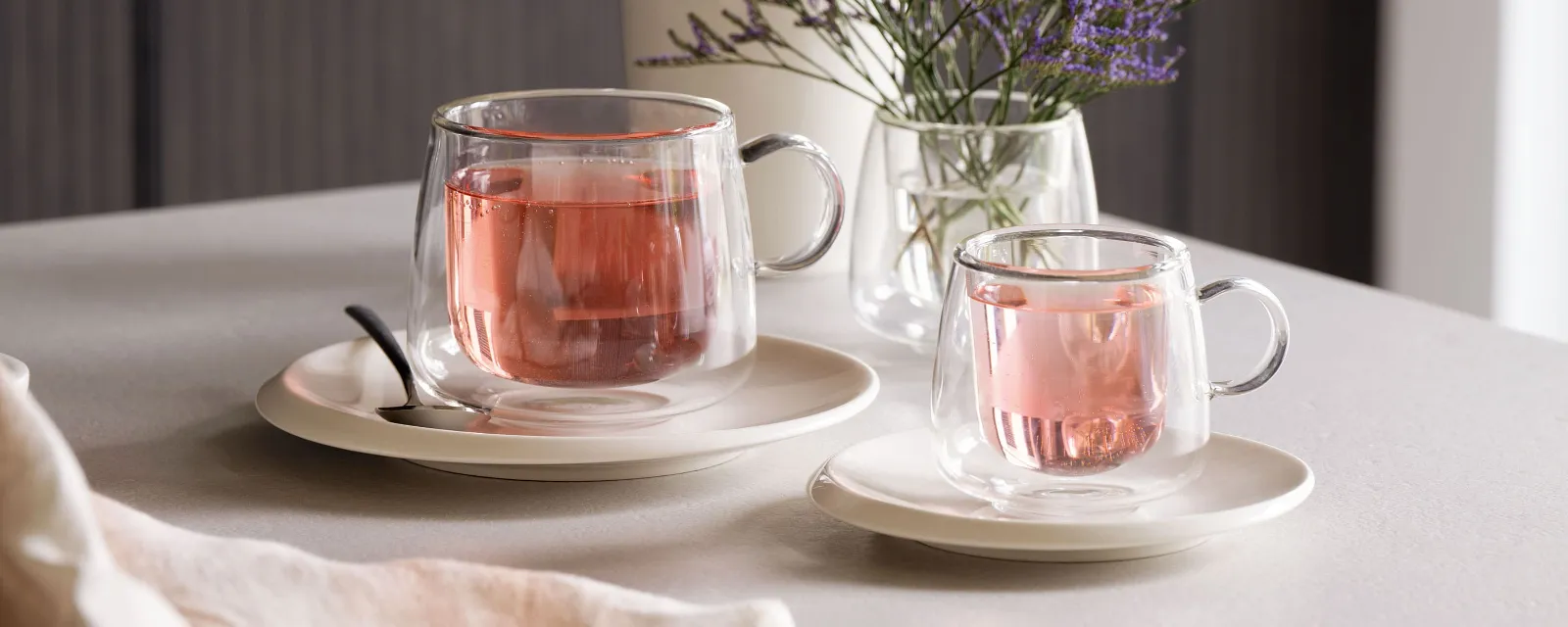 Artesano glass cups from Villeroy & Boch filled with pink tea, placed on saucers next to a spoon on a table that is decorated with a small vase of purple flowers.