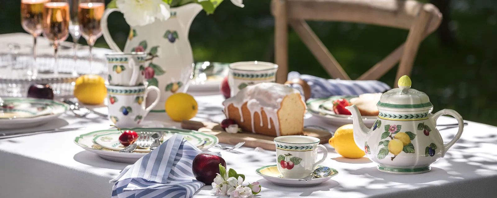 Outdoor table setting with floral French Garden porcelain, a teapot, rosé glasses and a cake. White peonies in a vase and fruit are arranged around the table.