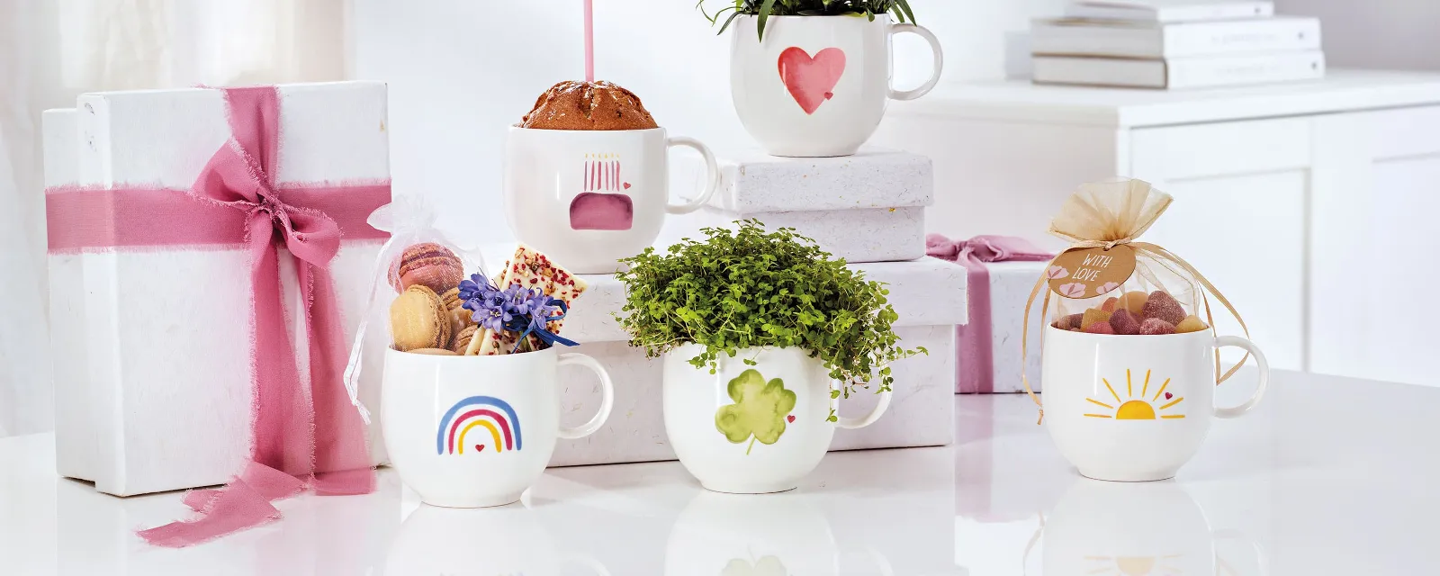 Five Villeroy & Boch white "With Love" cups, each with a different flower, sweet and gift pattern, on a white surface. In the background, two presents wrapped with pink bows complete the charming scene.