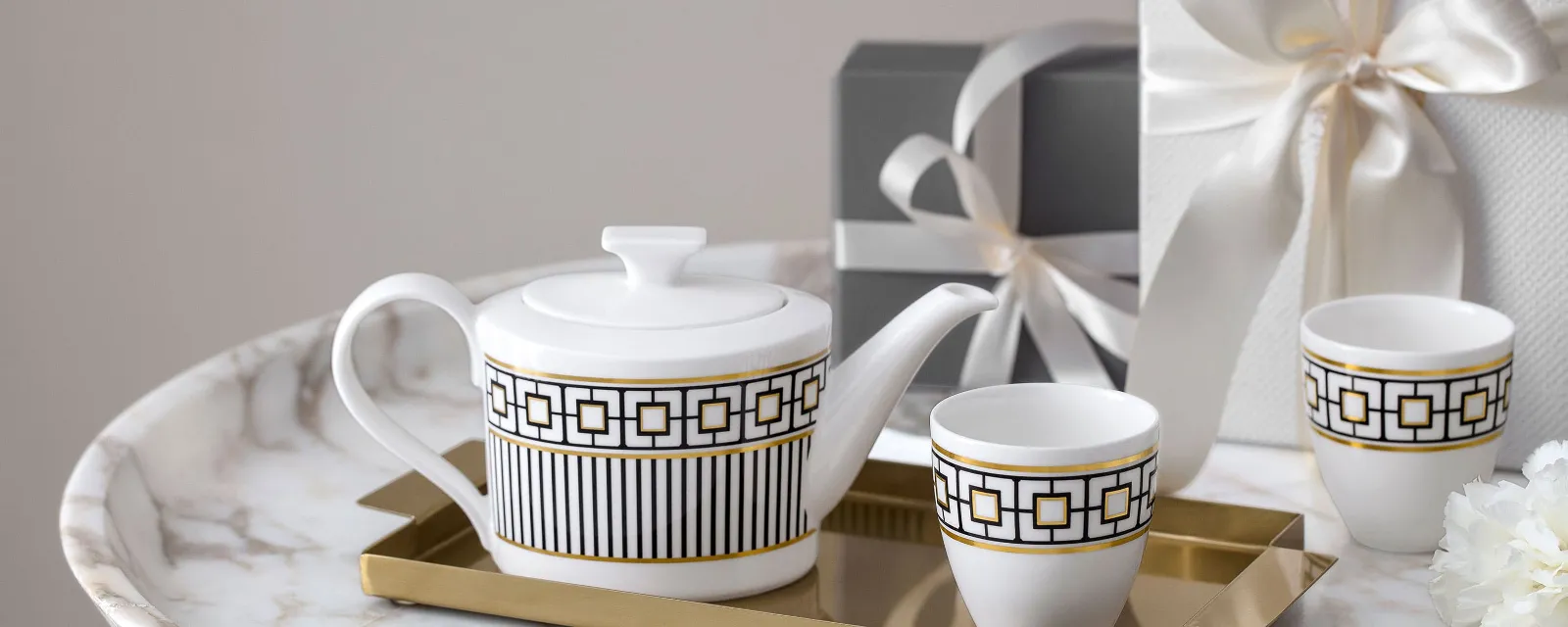 A white and gold patterned teapot and cups from MetroChic on a golden tray on a marble table. Two wrapped presents and a small white flower can be seen in the background.