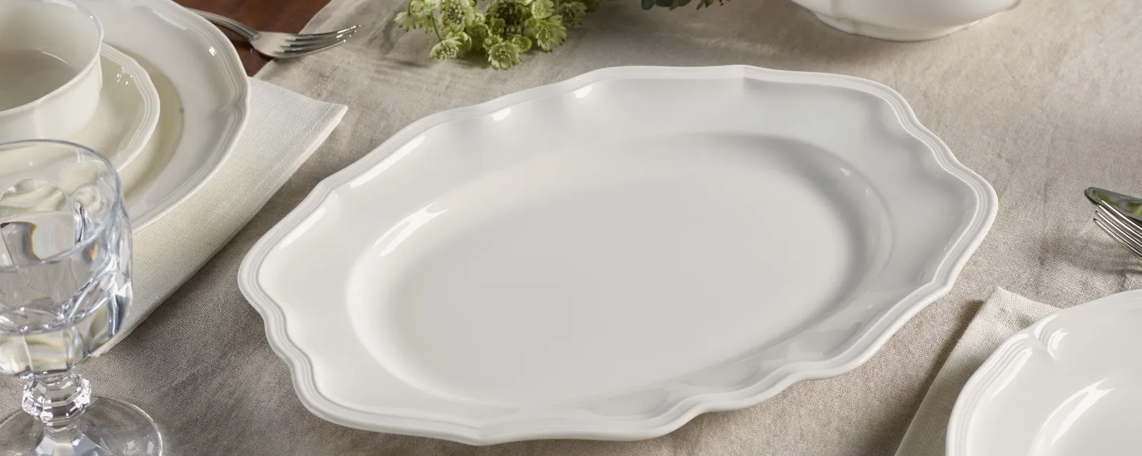 A white platter with scalloped edges is elegantly placed on a set dining table adorned with a tablecloth, surrounded by plates, glasses, cutlery, and bowls.