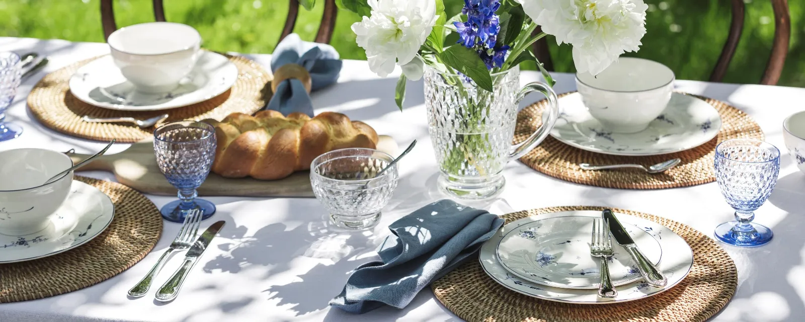 A garden table setting with Villeroy & Boch "Old Luxemburg" floral porcelain, wicker placemats, blue-tinted glass tableware, a crystal jug with blue and white flowers and a loaf of bread.