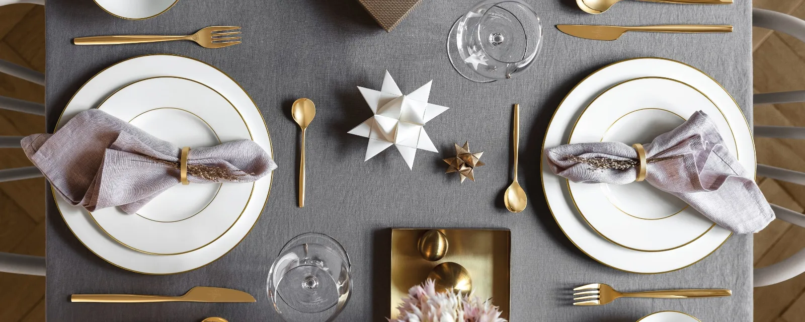 An elegant dining table setting with two Villeroy & Boch Anmut place settings, gold cutlery, lavender napkins, a gift box and a flower arrangement on a grey tablecloth.