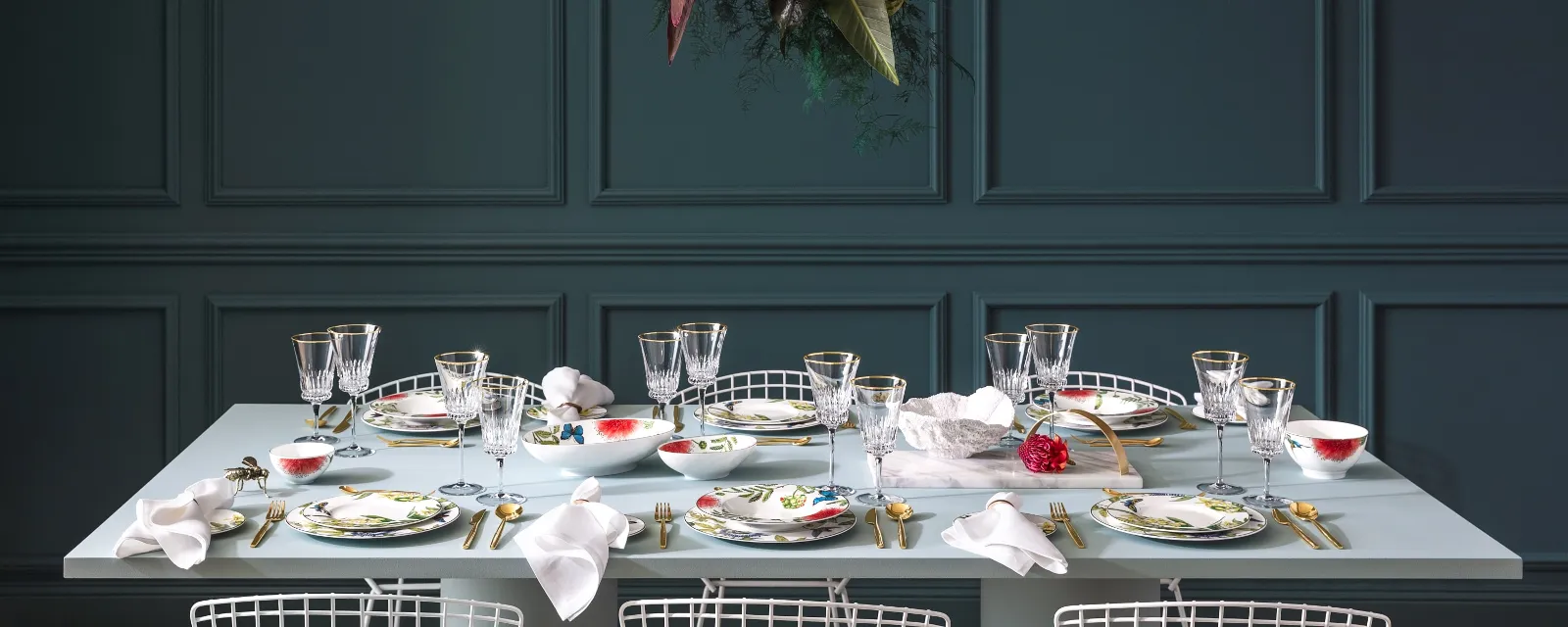 The Villeroy & Boch Amazonia dining table set consisting of glassware, gold cutlery and plates with floral patterns. A floral arrangement hangs above the dining table creating a beautiful contrast with the dark panelled wall.