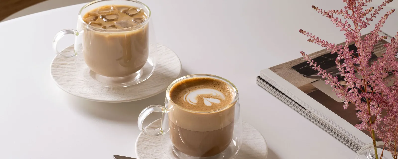 Two Villeroy & Boch Artesano glass tumblers on a table: one filled with iced coffee, the other with latte, plus a spoon, a stack of magazines and a pink flower in a vase.