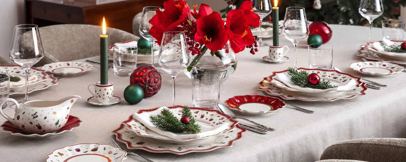 A festive dining table setting with elegant Toy's Delight crockery, lit candles, red flowers in a vase and Christmas decorations, with a decorated Christmas tree in the background.