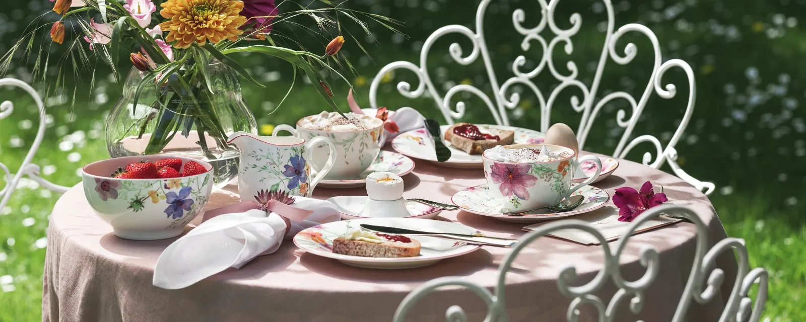 A Villeroy & Boch Mariefleur garden table set consisting of floral-patterned porcelain, a vase of colourful flowers and plates holding desserts and strawberries. Ornate white chairs surround the table against a background of lush green grass.