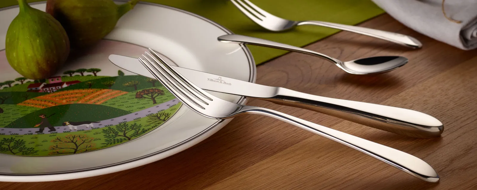 A white plate with a landscape pattern, two figs and a Sereno cutlery set arranged on a wooden table. 