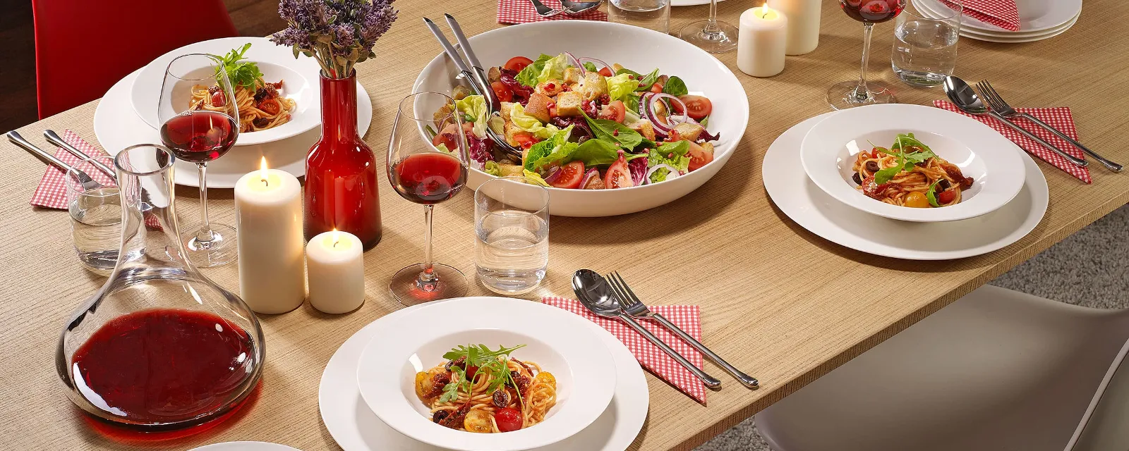 A dining table with For me plates filled with salad and pasta, with glasses of red wine, candles and a carafe of red wine.