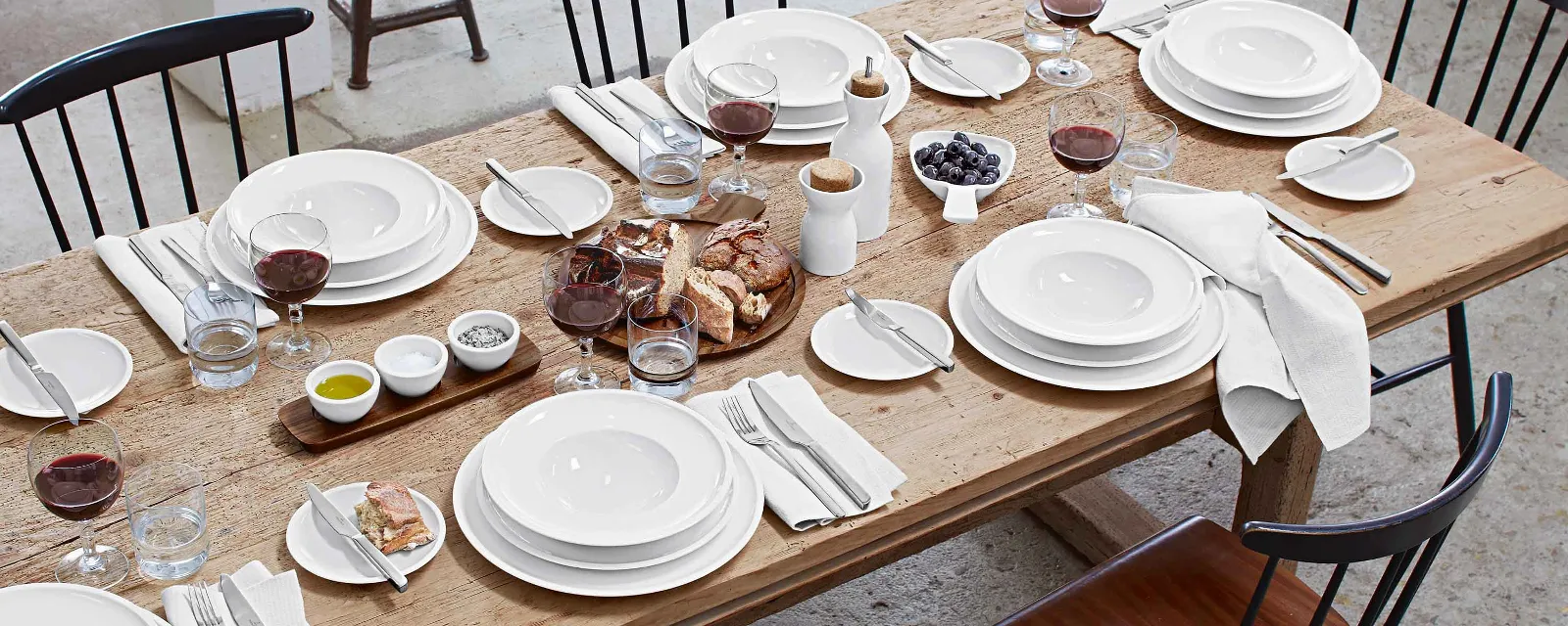 A wooden dining table for six people with white dinnerware, red wine glasses, napkins, bread, grapes, butter, salt, pepper, oil, and water glasses. Plates and flatware are placed at each place setting.