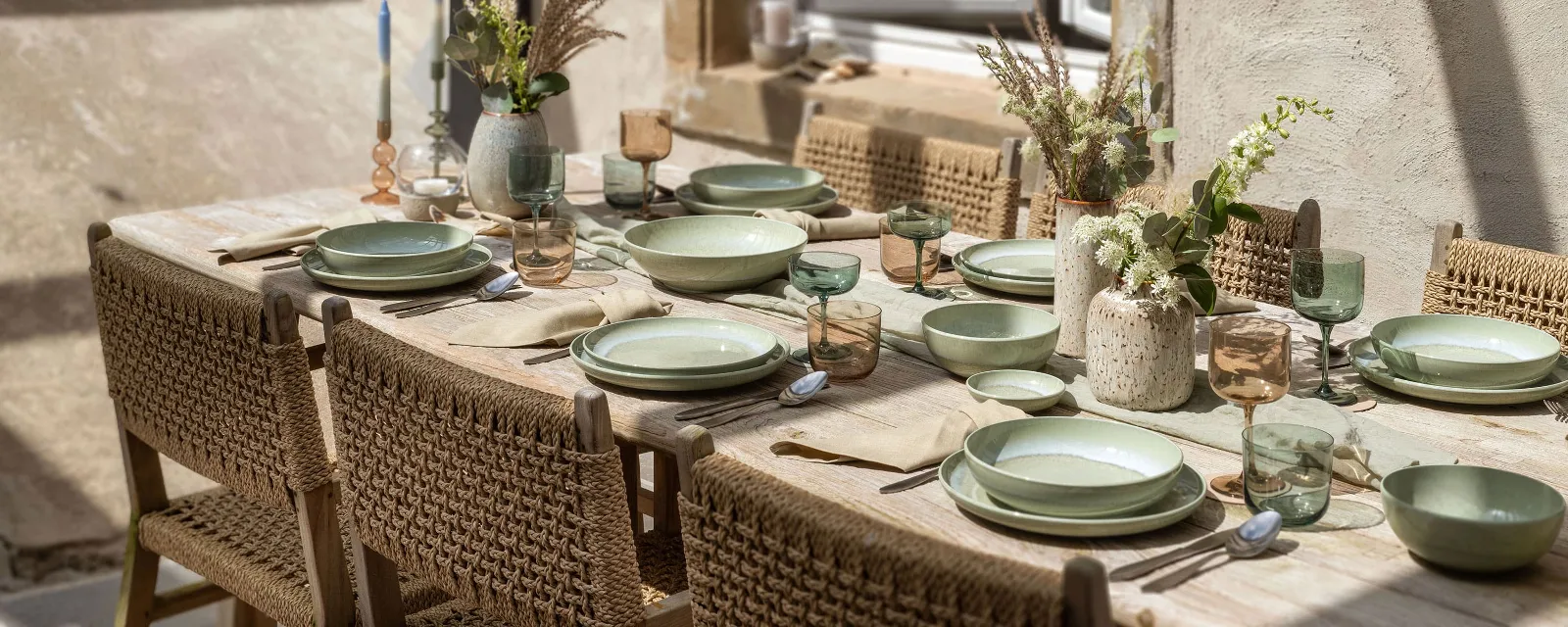 A summery dining table with green plates from the Perlemor collection, glasses and cutlery on a wooden surface. It is framed by wicker chairs and the centrepiece is adorned with decorative vases full of flowers.