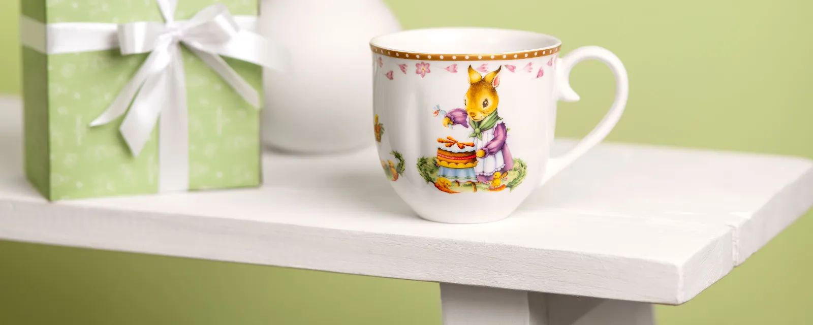 An Annual Easter Edition cup on a white table in front of a green gift box with a white bow and a vase of flowers