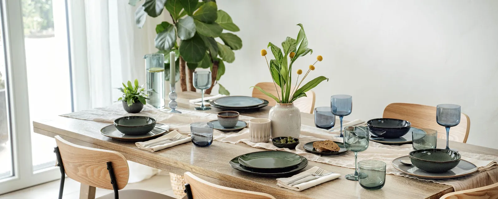 Dining table set with various items from the Lave collection by Villeroy & Boch The dining table set with various items from the Lave collection by Villeroy & Boch in green and grey, accentuated by blue glasses and napkins. A vase of yellow flowers and a small potted plant provide decoration, while a large plant stands by the window.